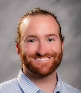 Headshot of a Caucasian man smiling. He has red hair and a beard. He is wearing a light blue collared shirt. 